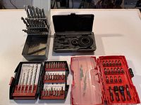 Overhead view of multiple drill bit sets, magnetic bit holder with screwdriver bits, and hole saw set in plastic case, with plastic bin lid underneath