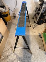 Full length top view of the folding steel saw horse showing the blue top surface with black non-slip strip and black folding legs.