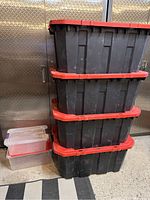 Stack of four large black Husky storage bins with red lids and two smaller clear storage bins next to them on the floor