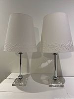 Front view of two identical lamps with shades on, showing the embroidered vine pattern on the bottom edge of the white shades and clear glass bases.