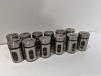 Front view of all 12 spice jars arranged in group showing metal sleeves and lids with dial tops