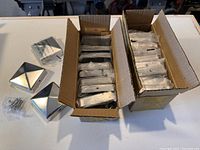 Box of new stainless steel fence post caps and plastic bags with screws on a white surface.