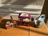 Three Limoges porcelain boxes including a fruit cart, watering can, and flower basket, shown together on a wooden surface with sunlight highlighting paint details and colors.