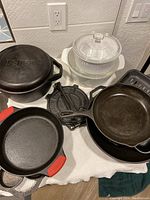 Photo showing multiple Lodge cast iron skillets and pans including one with red silicone handle covers, iron tortilla press, and two white casserole dishes with lids.