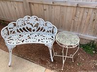 Photo showing full view of white cast iron garden bench with floral pattern and accompanying white metal side table with glass top outdoors next to a wooden fence.