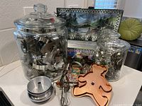 Full view of two large glass jars containing metal cookie cutters, copper-colored bear cookie cutter, vintage egg beater, and stackable aluminum baking pans on counter.