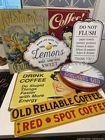 Photo of 6 vintage metal signs displayed on a table including coffee advertising signs, lemon sign, DO NOT FLUSH sign, and Gordon's Gin thermometer