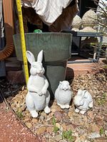 Photo showing large green ceramic garden pot behind three white ceramic rabbit figurines of varying sizes on a gravel bed.