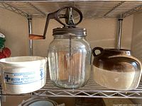 Full lot showing three antique pieces: glass hand crank butter churn and two pottery crocks on metal shelf.