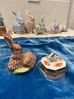 Figurine and trinket box displayed together on blue cloth with other porcelain rabbits in background