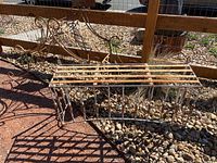 Two rustic iron garden decor pieces including a rectangular plant support with grid top and vertical spindles, and a curled iron arch support for vining plants, shown outdoors on gravel bed.