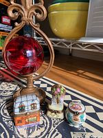 Set of three Limoges porcelain pieces: café box, floral pot, and reading chair figurine beside a red glass ball and metal stand