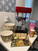 Front view of red Waring popcorn maker with two ceramic popcorn bowls, a ceramic popcorn mug, striped paper popcorn container, and two vinyl decorative movie theater signs.
