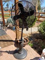 Full view of outdoor fan showing black metal fan head with blades, attached to a tall pole and weighted round base on patio.