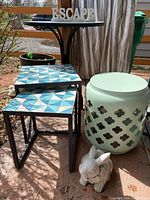 Photo showing two square metal tables with blue and white geometric tile tops, mint green metal garden stool with decorative cutouts, and a round mosaic blue tiled table in the background with wooden ESCAPE letters on top.