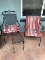 Front view of two black metal outdoor rocker chairs with red and white striped cushions and small metal mesh side table between them.