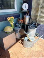 Wide angle showing galvanized watering cans, decorative ceramic figures, old metal box, and part of metal plant stand.