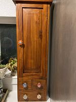 Front view of tall wood cabinet with door closed and ceramic flower-shaped handles on door and drawers.