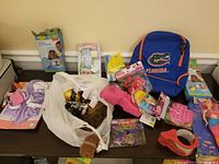 Photo showing an assortment of toys including a Florida Gators backpack, mermaid plush doll, R2-D2 toy figure, and various smaller toys and accessories laid out on a table.