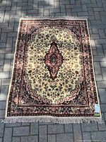 Area rug laid out flat on paved ground showing full rug with fringe at one end. The rug has a cream and red floral medallion pattern with a red floral border.