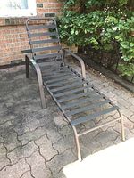 Outdoor metal chaise lounge chair on patio surface, showing full side angle and slatted seat design