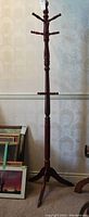 Full view of 71-inch tall wood coat rack stand with multiple hooks and tripod base in a room with wallpapered walls and framed pictures on the floor nearby.