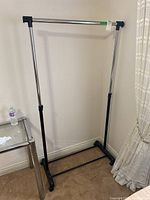 Front view of adjustable garment rack with chrome metal bars, black plastic connectors, and caster wheels on beige carpet near table and curtain.