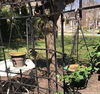 Outdoor photo showing two steel hanging planters each with a clay pot inside, hanging on a black metal frame against a garden background.