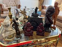 Overview of multiple figurines and folk art pieces on a glass table including Buddha statues, artisan carvings, brass candle holders, and miniature dragon figure