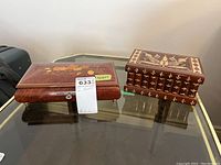Two lacquered wooden jewelry boxes on a glass top table, one with floral marquetry and one with geometric floral marquetry