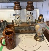Front view of the wooden stand holding two glass dispensers labeled 'Vodka' and 'Gin', two ceramic mugs, and the Kochamy oil lamp on a white surface.