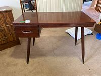 Front view of MCM wooden desk with one drawer and tapered legs on carpeted floor.
