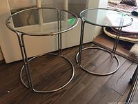 Pair of steel and glass nesting tables with tubular chrome finish frames and round glass tops, shown side by side on wooden floor.