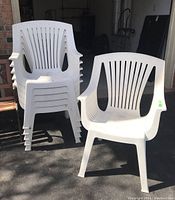 Six white resin outdoor chairs, stacked and one set out for display, outdoor setting.