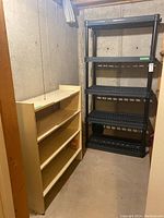 Photo showing three-tier cream colored wooden shelving unit and four-tier black plastic shelving unit side by side in basement corner.