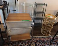 Photo of four furniture pieces: side table with fabric magazine holder and wooden top, white tiered stand, black shelf unit, and rattan shelf unit