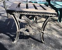 Side angle view of cast iron base of outdoor table highlighting dragonfly and leaf design and curved legs, with slatted wooden top.