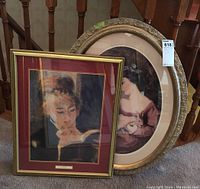 Two framed portrait prints leaning against a wooden stair railing. Left print is a Renoir art reproduction titled 'Young Woman Reading' in gold rectangular frame with maroon mat. Right print is an oval portrait of a woman holding a child and rabbit in an ornate oval frame.