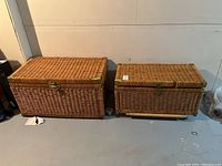 Two wicker storage trunks side by side on concrete floor against wall. Larger trunk on left, smaller trunk on right, both on wooden bases with metal latches and visible wear.