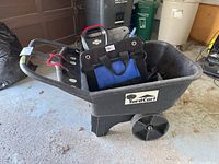Black plastic Yard Cart with two wheels, HardCart label on side, containing blue and black bags, red cables visible inside. Several trash bins and other garage items in background.