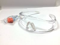 Clear shooting glasses with white temples and nose cushion next to a sealed bag of orange foam ear plugs.
