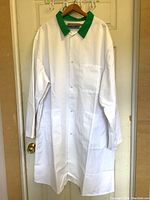 Full front view of the new heavy duty white butcher coat with green collar hanging on hanger against a door.