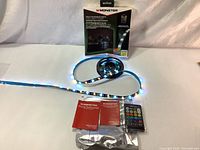 Complete lot including Monster Basics LED light strip partially unrolled on black reel with blue and white LEDs lit, remote control, USB cable, manuals, and original box visible in background.
