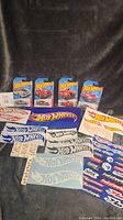 Full view of five Hot Wheels cars in original packaging displayed upright with multiple Hot Wheels flame logo and blue logo sticker sheets spread below them on a dark background.