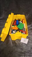 Yellow Lego storage tote with lid open showing assorted Lego pieces inside including bricks and wheels