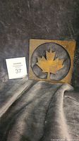 Square metal artwork featuring a maple leaf cutout, placed against a dark fabric background.