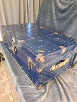 Side and top angled view of a blue Travelgard trunk showing metal corner protectors, locks, handles, and surface wear.