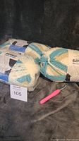Three skeins of Bernat Home Bundle yarn in white, blue, and beige colors with a pink crochet hook resting beside them on a dark fabric surface.
