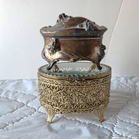 Two antique jewelry boxes stacked; one is a small spelter piece with patina, the other an ornate gold-toned filigree metal box with glass top