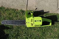 Top view of a green Poulan electric chainsaw lying on grass and concrete edge, showing the full chainsaw including the chain, blade, body, and handles.
